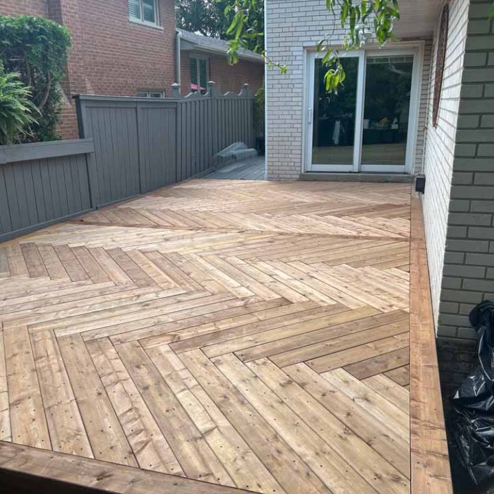 Close-up view of a nearly completed wooden deck installation, showcasing a herringbone pattern with varying tones of wood, enhancing the aesthetic appeal beside a residential home.