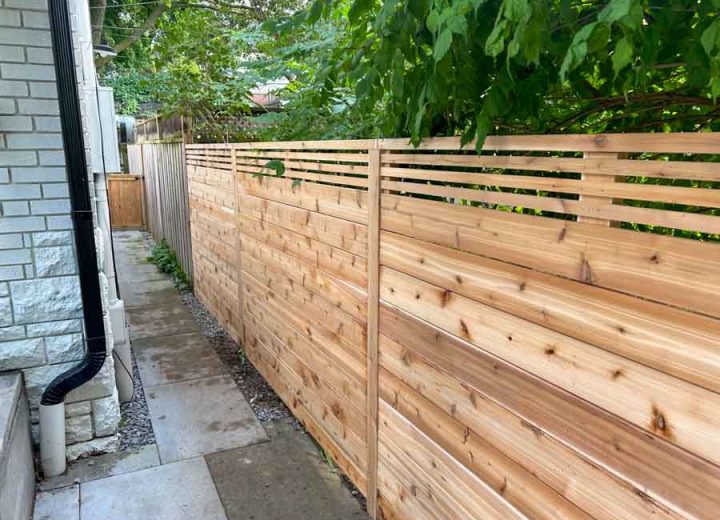 A contemporary wooden fence with horizontal slats along a narrow stone pathway, bordered by lush greenery and a residential building.