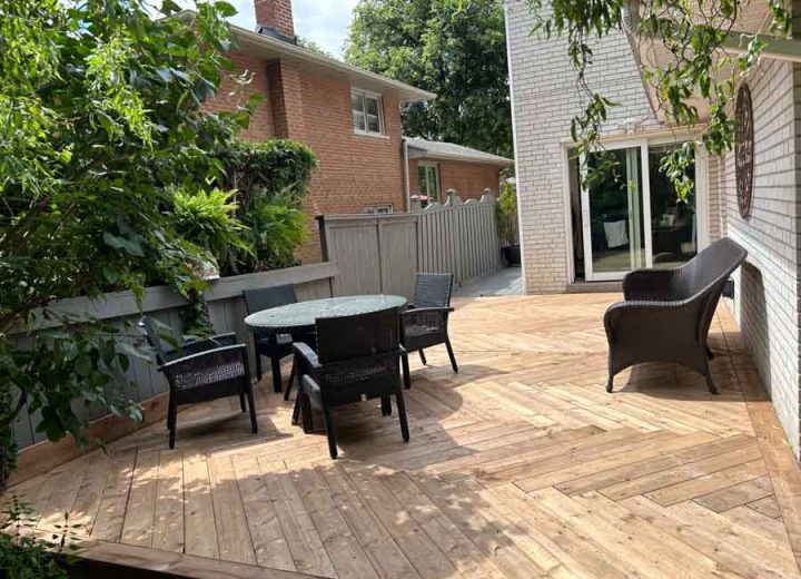 A cozy patio deck featuring a parquet pattern, furnished with a black dining set and a single lounge chair, enclosed by greenery and a grey privacy fence.